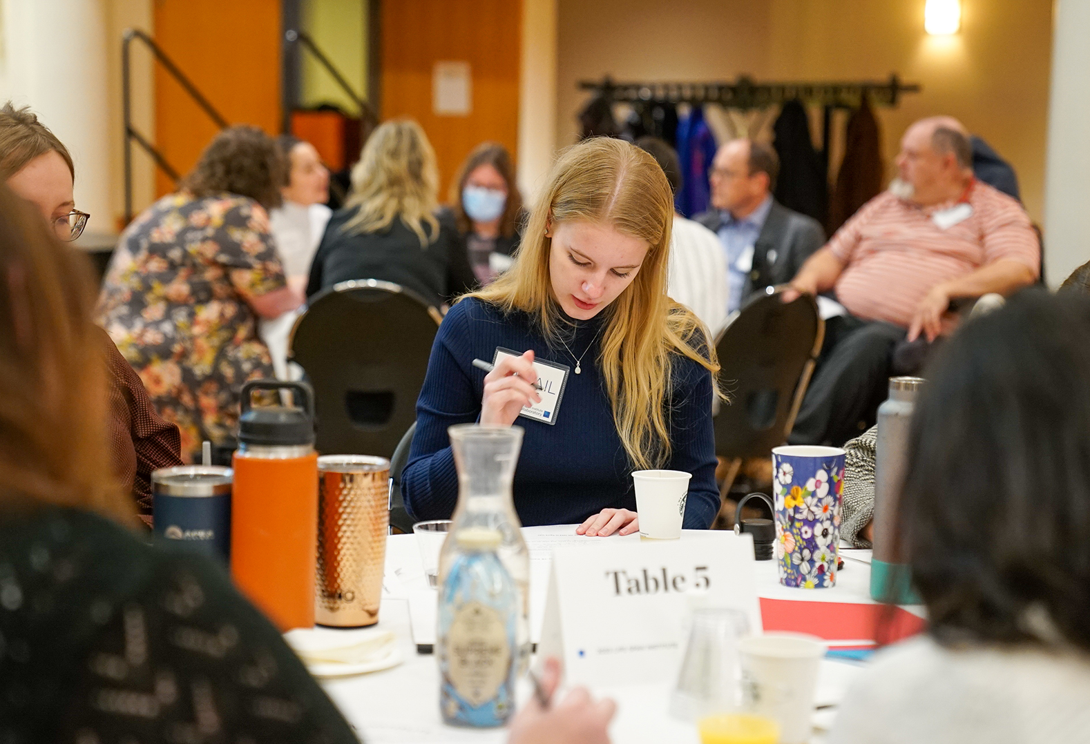 A woman with long blonde hair is in focus and surrounded by people at her table and other tables. She is holding a pen and looking down at a piece of paper. 