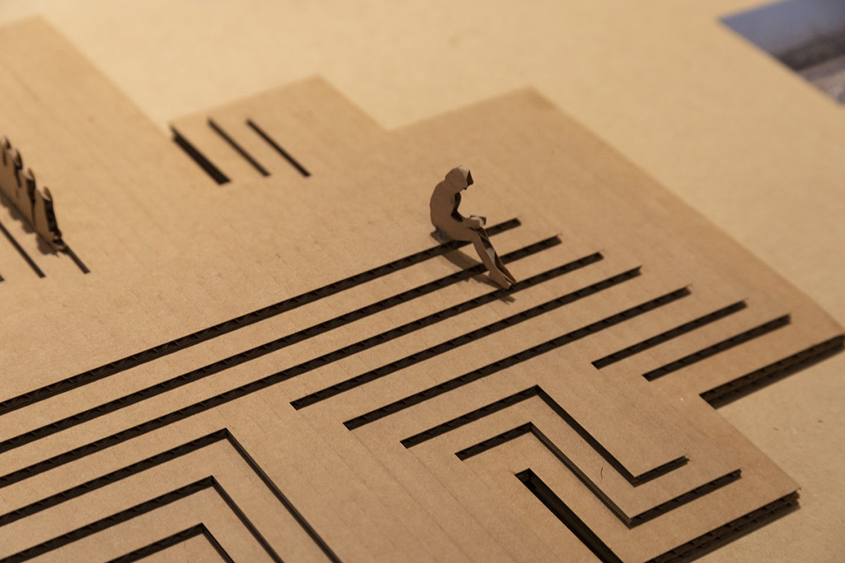 Close-up of a cardboard architectural or design model featuring a maze-like pattern of laser-cut grooves. A small paper figure sits on the edge of one of the cut channels, emphasizing scale. The model is displayed on a tabletop under warm lighting, with portions of additional model elements visible in the background.