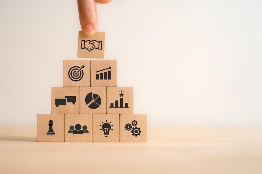 A hand arranges blocks that represent different aspects of business relationships, from ideas to profitability to manufacturing to sales.