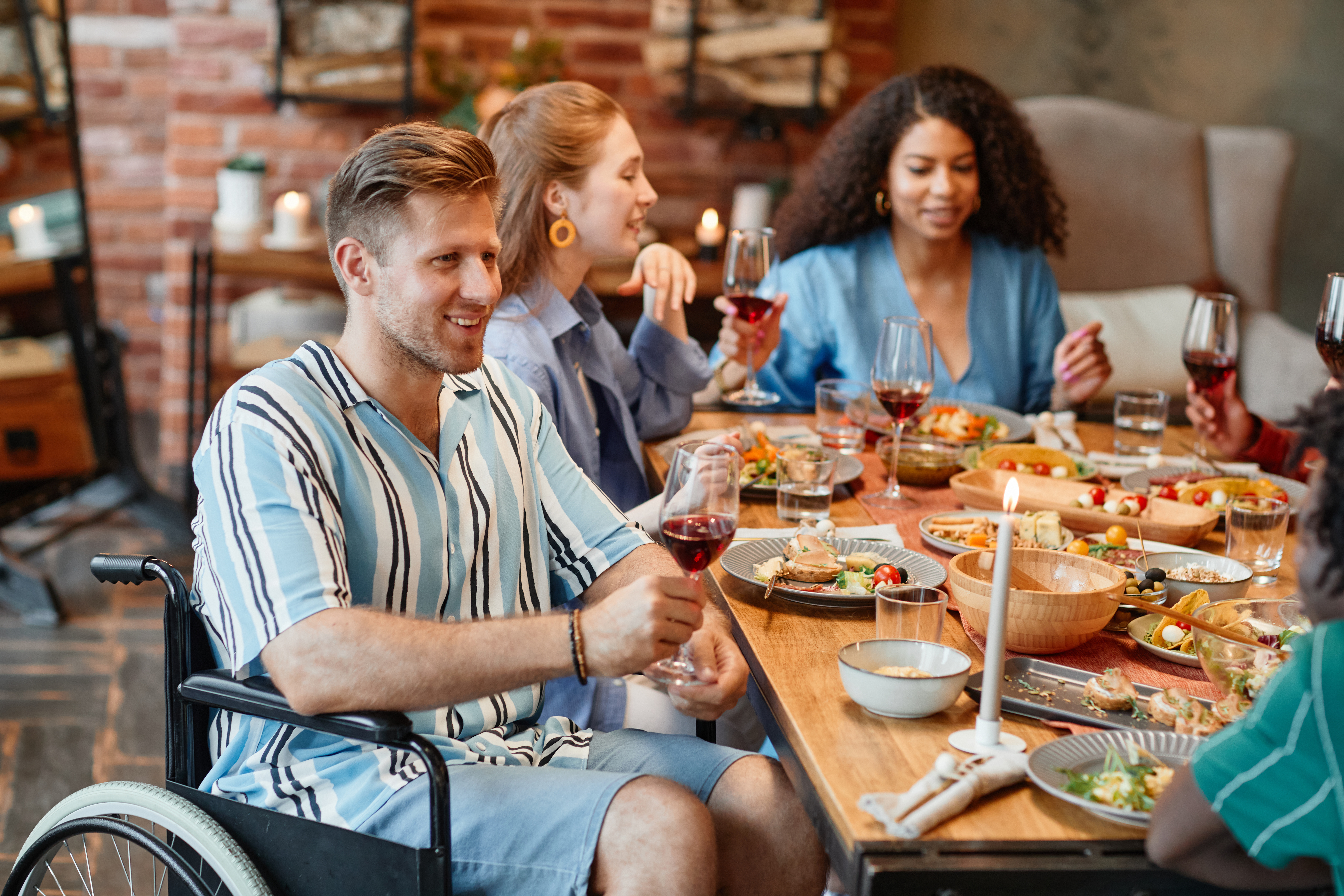 "A man with a striped shirt sits in a wheelchair at a table with friends eating dinner together"