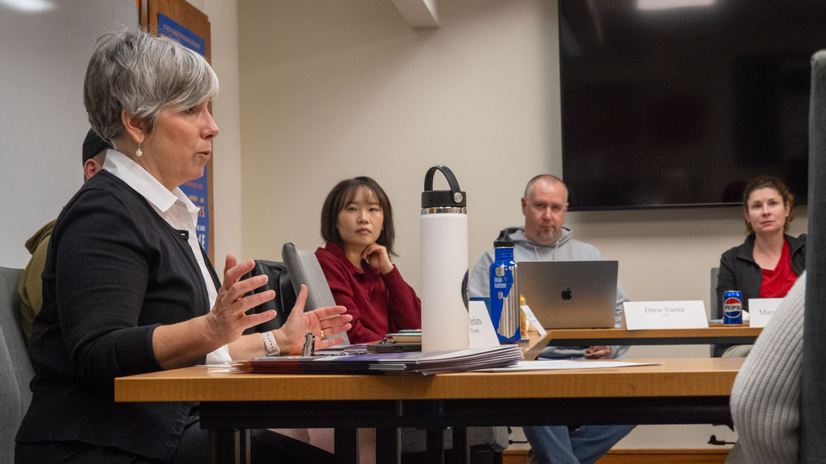 Woman sits at conference table speaking to others around table.