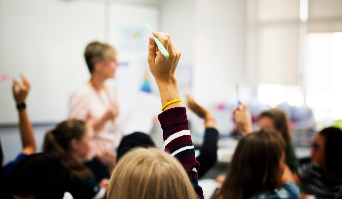 An image of a classroom full of students raising their hands while a teacher watches from the front of the room.