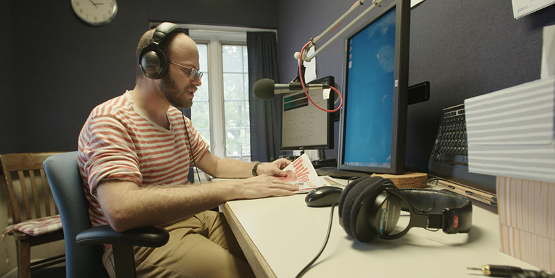 Volunteer reader Julian Silverman sits at the microphone in an Audio-Reader studio wearing headphones while reading the Hy-Vee ad.