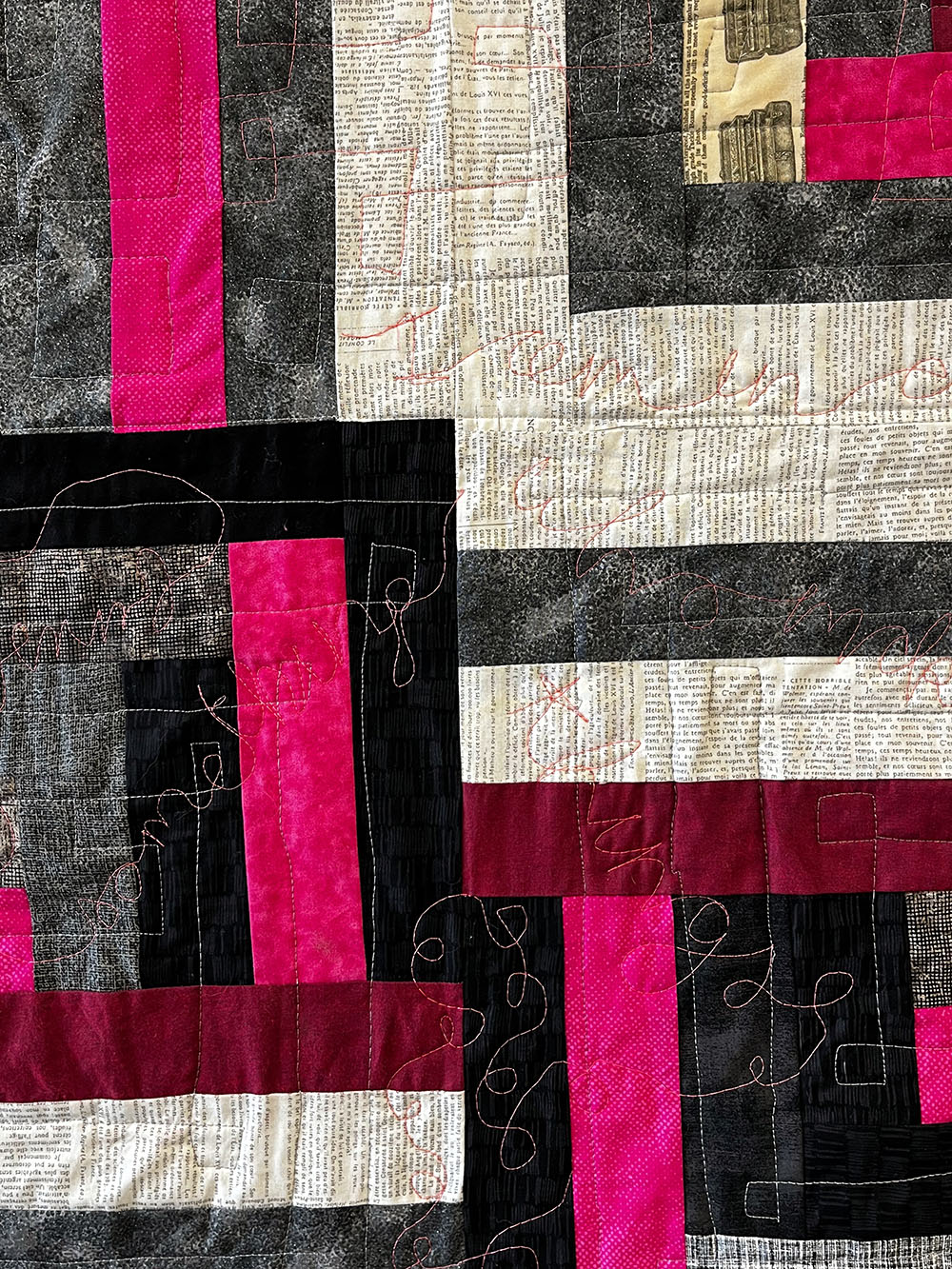 Quilt with blocks of fuschia, grey, black and gold-embroidered text.
