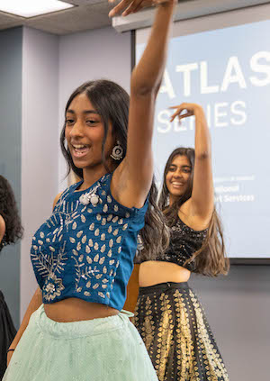 Two student dancers extend right arms toward ceiling in classroom with ATLAS series logo on screen projector.