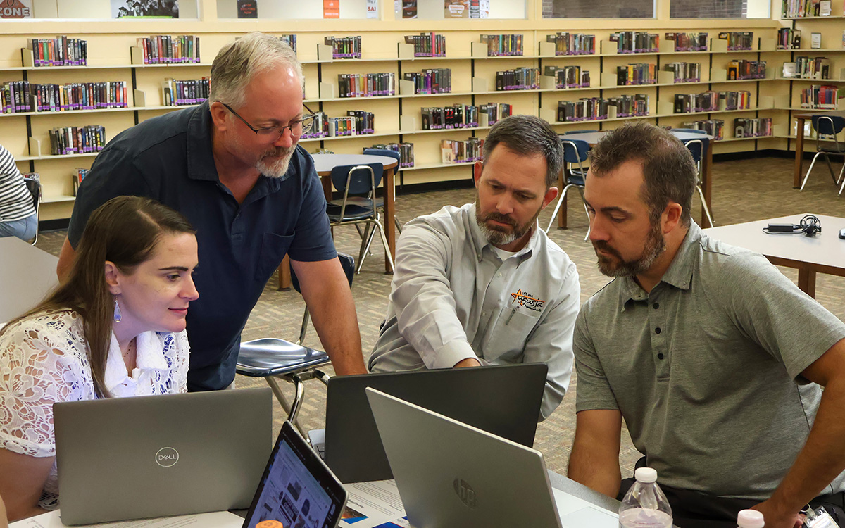 Four educators collaborate around laptops in a library during a professional development session. One person is standing and pointing at a screen while the others focus attentively, highlighting teamwork and engagement in a learning environment.