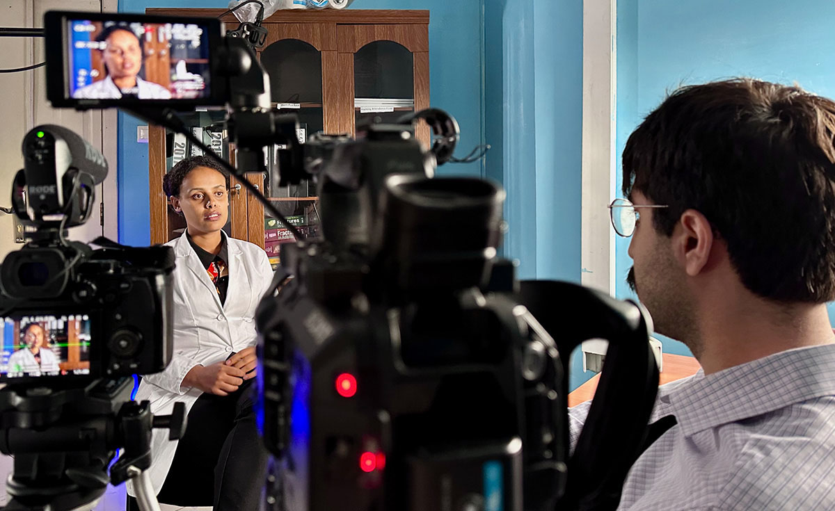 Man standing at right, operating a camera, interviewing woman seated at left.