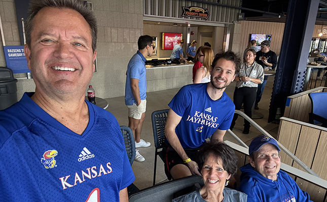 Two people in Kansas Jayhawks jerseys taking a selfie, with two more seated behind them, all smiling at a sports stadium.
