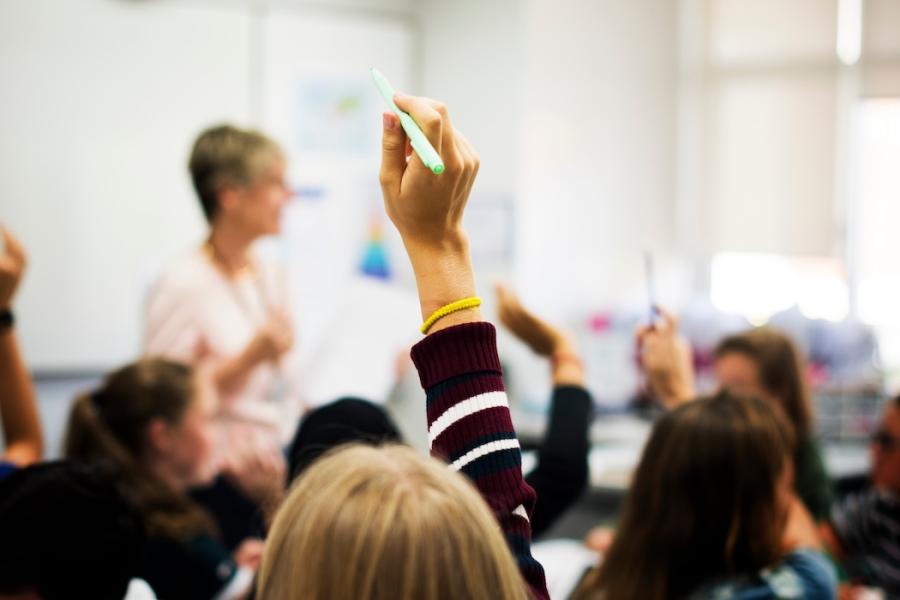 An image of a classroom full of students raising their hands while a teacher watches from the front of the room.