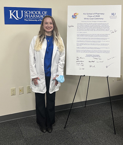 A person stands smiling beside a large poster on an easel. The poster is labeled "KU School of Pharmacy Class of 2026 White Coat Ceremony" and includes signatures. The individual wears a white coat and blue shirt. In the background, a sign reads "KU School of Pharmacy The University of Kansas."
