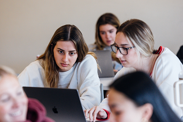 Two girls collaborate together on a laptop in a classroom setting.