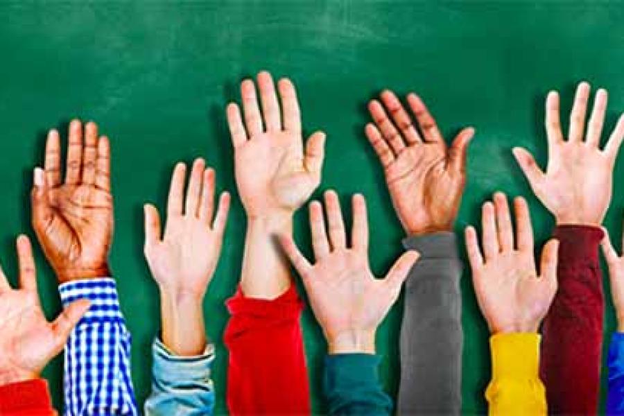 An image of several young students raising their hands in front of a chalkboard.