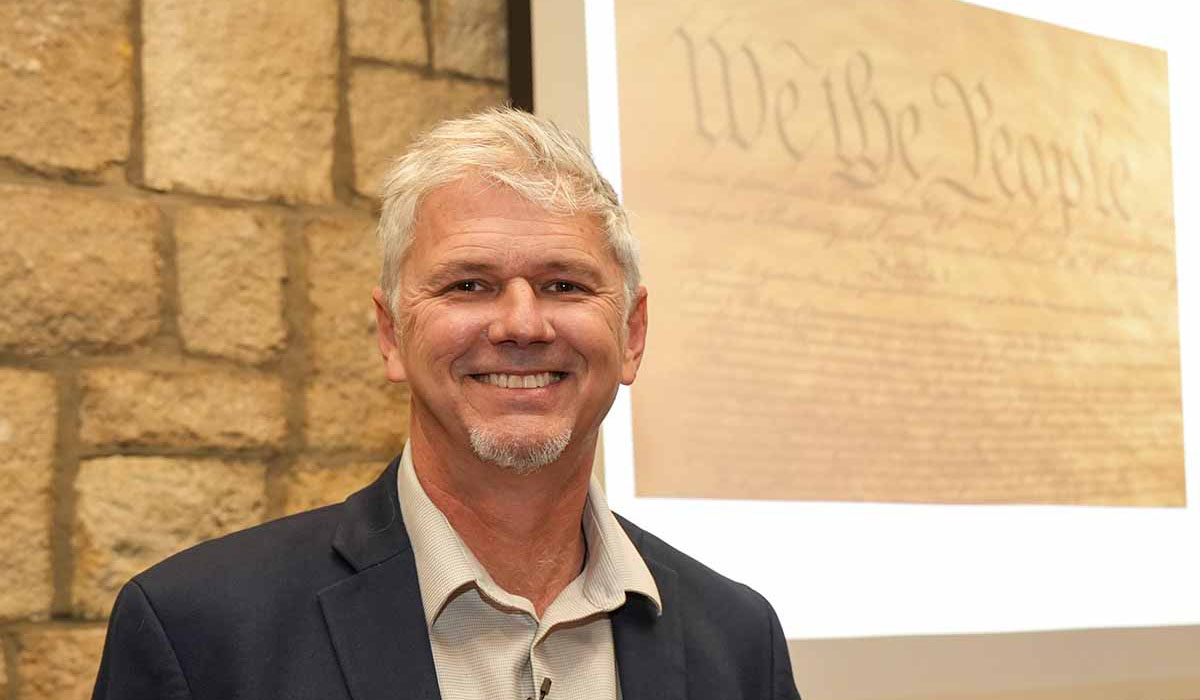 KU researcher Chris Koliba standing in front of background with "We the People ..."