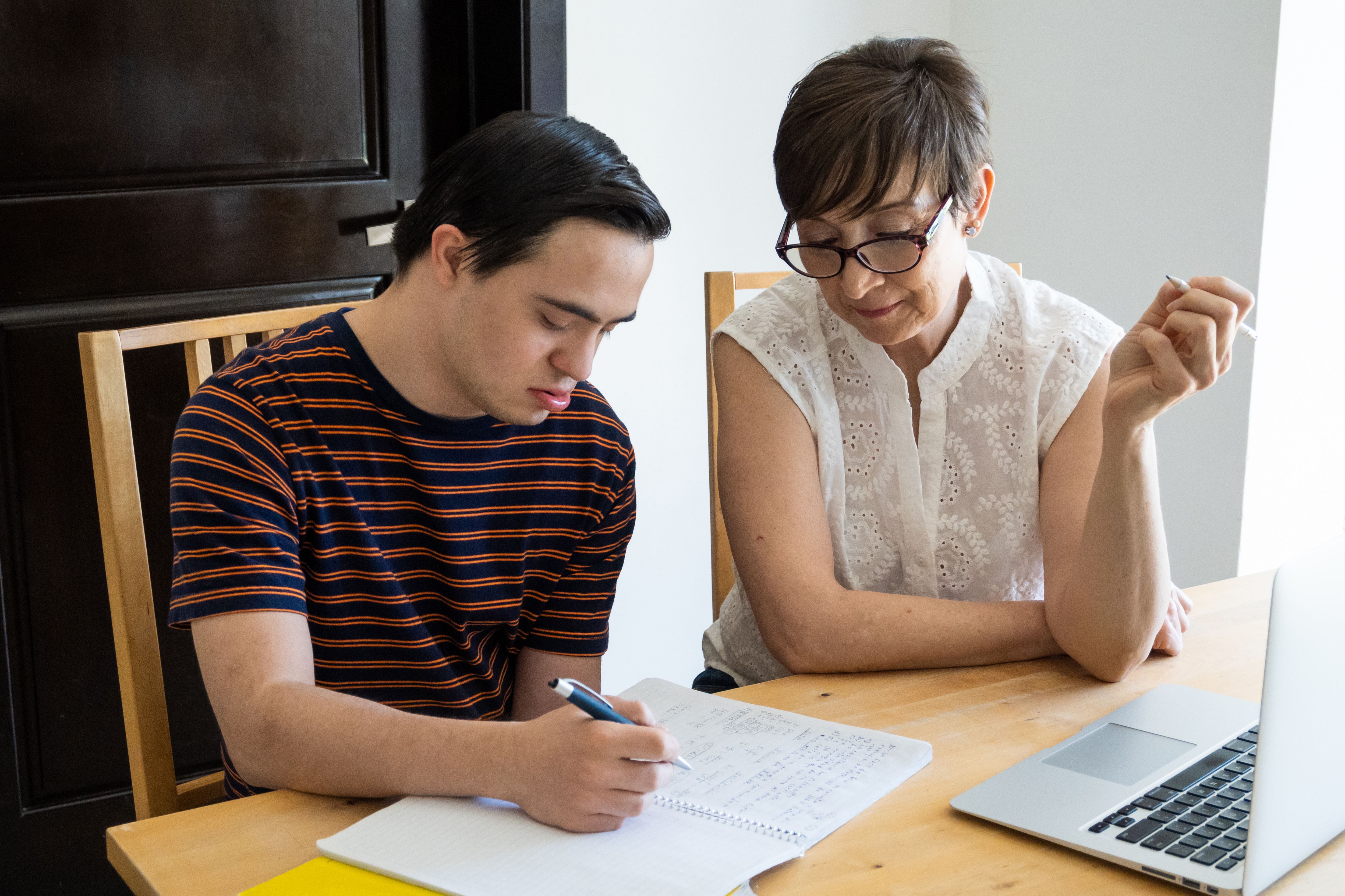 "A woman sits next to a young man at a table with an open laptop as he writes in a paper notebook"