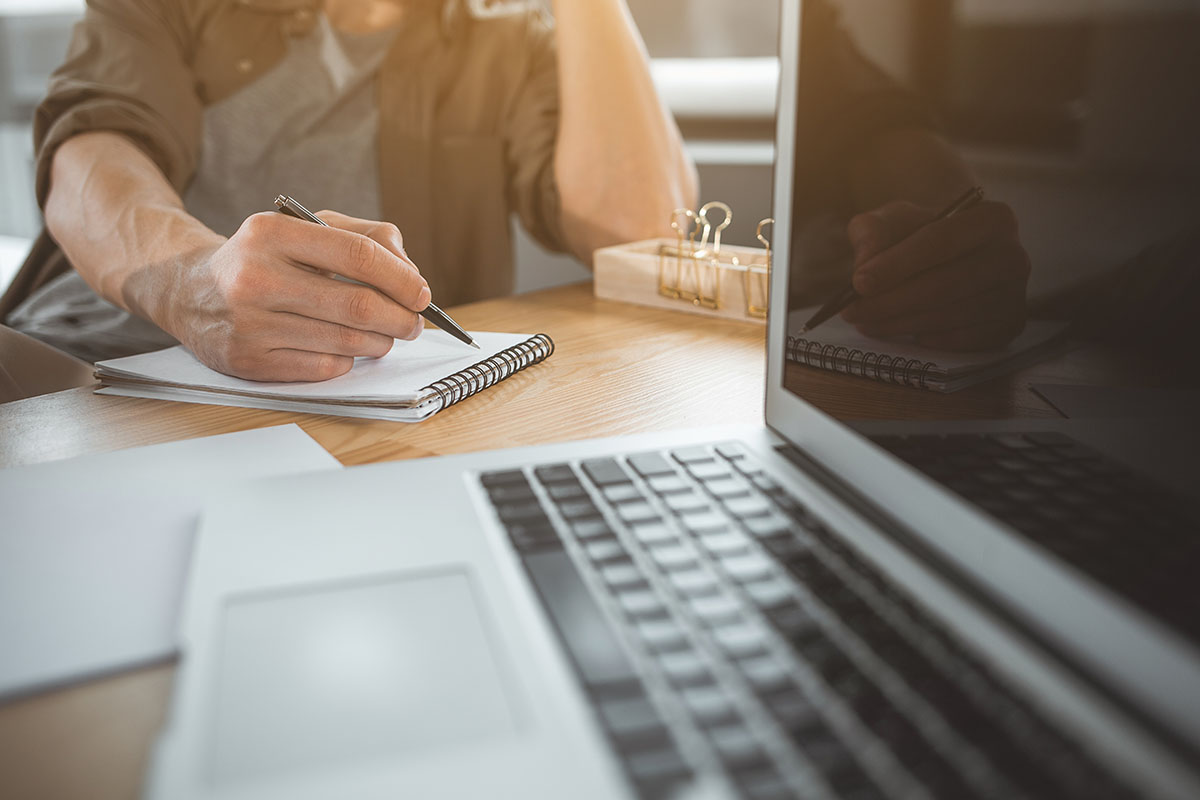 A man taking notes by hand, with a laptop sitting nearby