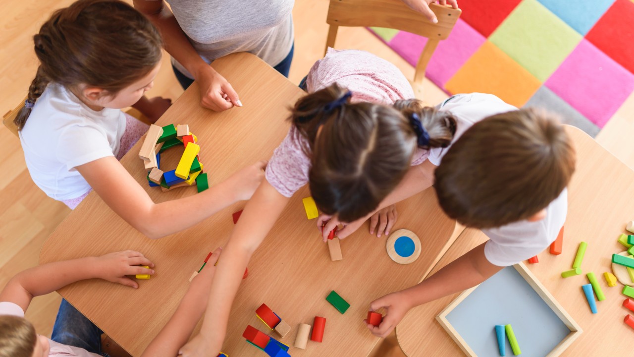 Three children play with blocks at a table in a playroom